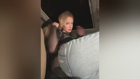 Media: Video of a blonde woman in a black top and light jeans, seated in a car, looking concerned; blurred background, dim lighting.