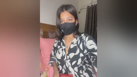 Media: Video of a young woman with medium brown skin, straight black hair, and a black face mask, wearing a black and white floral blouse, seated on a pink couch.