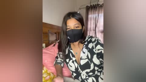 Media: A video of a young woman with medium skin tone, wearing a black face mask, black and white patterned blouse, and sitting on a bed with pink and yellow pillows, in a dimly lit room.