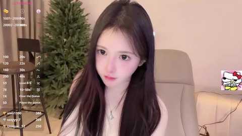 Media: Video of a young Asian woman with long black hair, pale skin, and a slender build, seated indoors near a Christmas tree, wearing a white top.