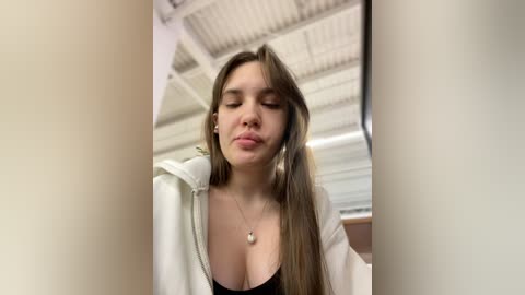 Media: Video of a young Caucasian woman with long brown hair, wearing a black top and a white jacket, standing in a modern, industrial-looking room with white walls and a corrugated ceiling.