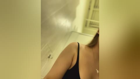 Media: Video of a woman's bare back in a dimly lit hallway with smoke wafting from an open door on the left. She wears a black spaghetti-strap tank top, and her light brown hair is pulled back. The background features off-white walls and a staircase.