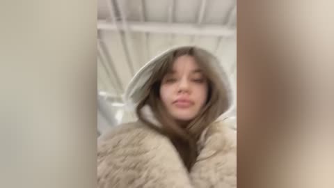 Media: Video of a young woman with straight, shoulder-length brown hair, wearing a beige fur-lined hooded jacket, making a pouty face. Background is a blurred, industrial interior with white ceilings and metallic beams.