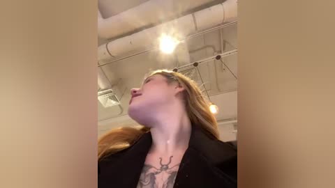 Media: Video of a fair-skinned woman with long, wavy blonde hair, wearing a black blazer over a floral-patterned top, with visible tattoos on her neck and chest. She has her head tilted back, gazing upward. The background features industrial ceiling pipes and a spotlight.