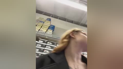 Media: Video of a blonde woman in a black blazer, leaning back, with a blurred office background featuring filing cabinets and a white ceiling.