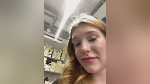 Media: Video of a young, fair-skinned woman with long, wavy auburn hair, smiling slightly, taken in a sterile, well-lit room with yellow boxes and metallic ceiling.