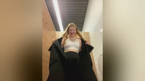 Media: Video of a smiling, fair-skinned, blonde woman in a white crop top and black pants, standing in a narrow hallway with white brick walls and a wooden door, holding a black jacket.