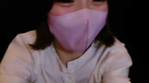 Media: A close-up video of a person wearing a pink surgical mask and a light-colored shirt against a dark background. The image is slightly blurred, focusing on the mask and upper body.