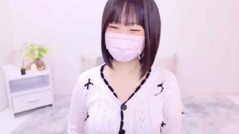 Media: Video of an East Asian woman with short black hair, wearing a white lace-up cardigan, white mask, and sitting in a minimalist room with white walls and a small white table with a vase.