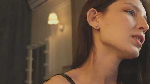 Media: Video of a young woman with straight brown hair, fair skin, wearing a black top, standing in a dimly lit room with a warm, yellow wall sconce and wooden staircase in the background.