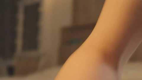 Media: A close-up video of a light-skinned person's leg, blurred background showing a beige wall with a window and green curtains. The image has a soft, warm tone.