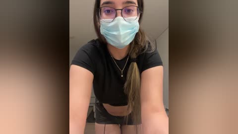 Media: A video of a young woman with light skin and long dark hair in a ponytail, wearing a blue surgical mask, black-rimmed glasses, and a black crop top, leaning forward.