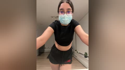 Media: Video of a young woman with fair skin, wearing a blue surgical mask, black-rimmed glasses, and a black crop top with black shorts. She stands in a minimalist room with light wooden floors and white walls.