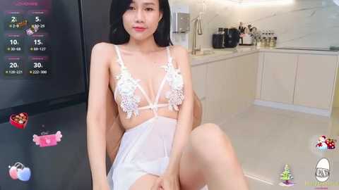 Media: Video of an East Asian woman with long black hair, wearing a white lace lingerie bodysuit, sitting in a modern kitchen with a digital display and kitchen utensils in the background.