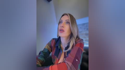 Media: Video of a blonde woman with fair skin, wearing a colorful plaid shirt, looking thoughtful, in a dimly lit room with a blurry background.