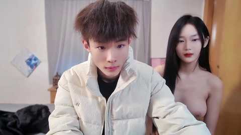 Media: Video of an Asian man with messy hair wearing a light jacket, and a nude Asian woman with long black hair and red lipstick, in a modern, brightly-lit room.