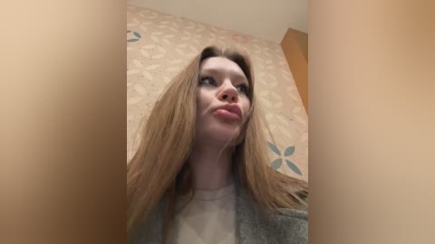 Media: A video of a young woman with long, straight brown hair and fair skin, making a pouty face, wearing a gray cardigan over a white shirt. The background features a beige floral-patterned wallpaper.