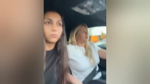 Media: Video of two women in a car; one with long dark hair, the other with blonde hair, wearing white tops, sitting side by side, blurry background.