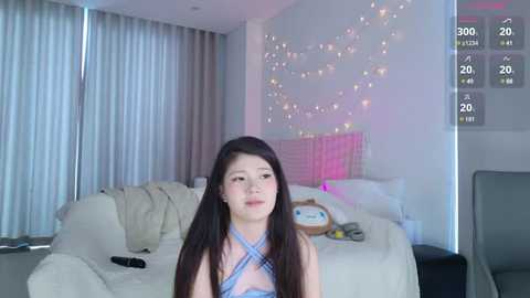 Media: Video of an East Asian woman with long black hair, wearing a blue plaid halter top, in a modern, cozy bedroom with white walls and fairy lights.