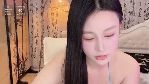 Media: A video of an Asian woman with long black hair, fair skin, and delicate facial features, wearing a light-colored top, sitting on a bed with white linens and a patterned wallpaper background.