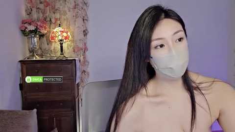 Media: Video of an East Asian woman with long black hair, wearing a surgical mask, seated in a room with a floral-patterned curtain, a dark wooden dresser, and a pink lamp.