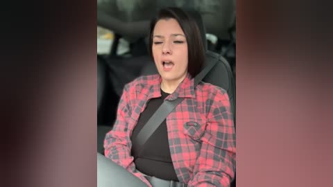 Video of a young woman with shoulder-length dark hair, wearing a red and black plaid shirt, grey seatbelt, and black top, laughing with mouth open, in a car.