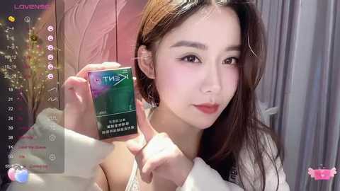 Media: A video of an East Asian woman with fair skin and long brown hair, wearing a white top, holding a green and white \"T-MAX\" beauty cream box, in a pink room with a calendar overlay.