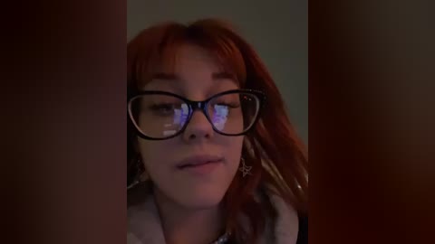 Media: A video of a young woman with fair skin, red hair, and large black-framed glasses, set against a dark, blurry background, wearing a gray coat.