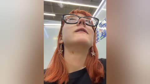 Media: Video of a woman with fair skin, shoulder-length red hair, and black-framed glasses, wearing star-shaped earrings, standing in an indoor setting with fluorescent lights and a colorful poster in the background.