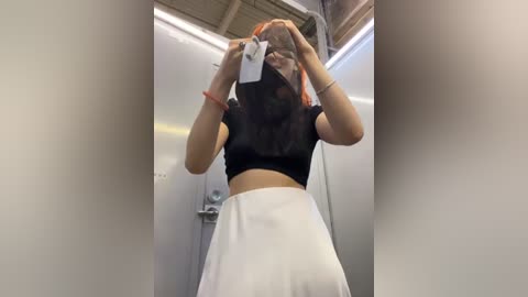 Media: A video of an Asian woman with shoulder-length black hair, wearing a black crop top and white high-waisted skirt, taking a selfie in a public restroom with a metal door and fluorescent lighting.