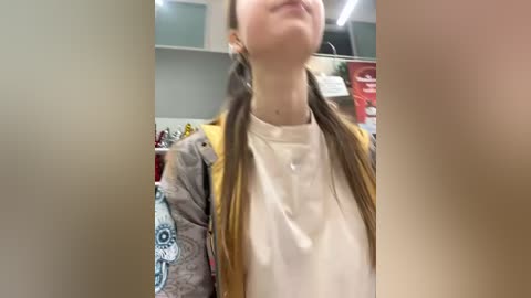 Media: Video of a young woman with fair skin, long brown hair in pigtails, wearing a beige shirt and a yellow jacket, standing in a brightly lit indoor setting with shelves and products in the background.