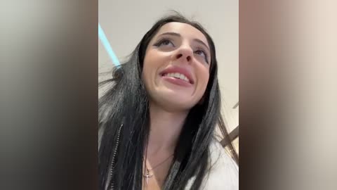 Media: Video of a smiling woman with long black hair, wearing a white top, in a blurred, indoor setting with beige walls.