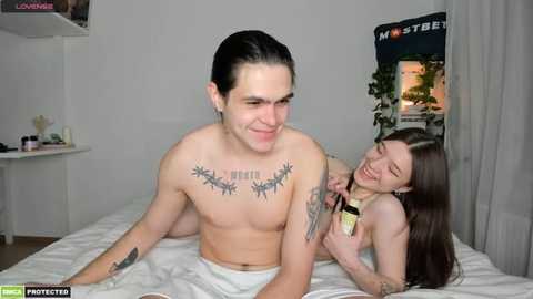 Media: Video of a shirtless man with long dark hair and multiple tattoos, smiling while embracing a topless woman with long brown hair, both on a bed in a minimalist bedroom.