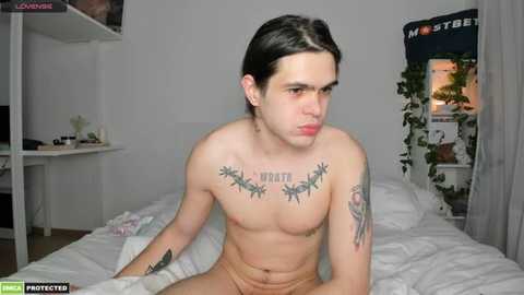 Media: Video of a shirtless young man with fair skin, long dark hair, tattoos including \"MOTHER\" on chest, sitting on a bed in a minimalist bedroom.