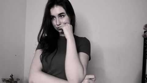 Media: Black-and-white video of a young woman with long dark hair, wearing a short-sleeved t-shirt, leaning against a plain wall, her elbow on her chin, expression contemplative.