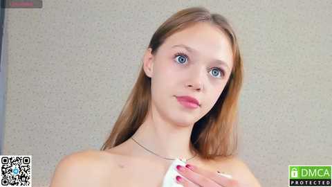 Media: Video of a young Caucasian woman with fair skin, blue eyes, and straight brown hair, wearing a white top, holding a white cloth. She stands against a beige textured background, with a QR code and \"DMCA Protection\" logo visible.