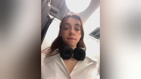 Media: Video of a young woman with long brown hair wearing a white shirt and black headphones, taken from a low angle, with a blurry background and a round light source above.