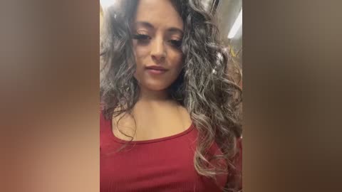 Media: A video of a young woman with long, curly, dark hair and light brown skin, wearing a red top, indoors.