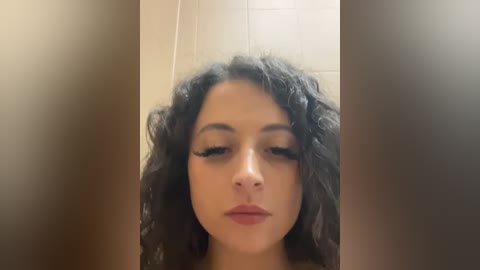 Media: A video of a young woman with curly black hair, fair skin, and full lips, wearing dark eyeliner and mascara, looking straight ahead. Background shows a beige tiled wall.