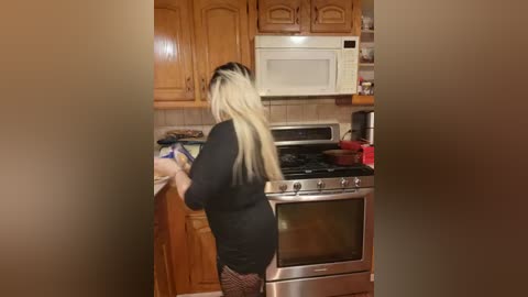 Media: Video of a blonde woman with long hair, wearing a black dress and fishnet stockings, baking in a kitchen with wooden cabinets, stainless steel appliances, and a red bowl on the counter.