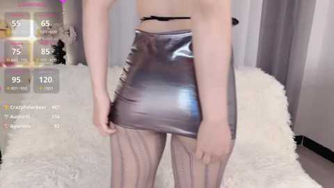 Media: Video of a slender, light-skinned woman with a small waist and long legs, wearing a shiny, metallic silver mini-skirt and sheer, striped tights. She stands in a bedroom with white fur carpet, gray curtains, and a digital screen overlay showing stats.