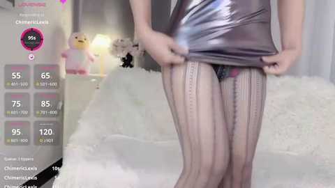 Media: Video of a person wearing a shiny silver mini dress and black fishnet stockings, standing in a bedroom with plush toys and a fluffy white bed.