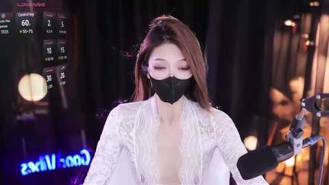 Media: Video of an Asian woman with long brown hair, wearing a black face mask and white lace dress, speaking into a microphone, against a dark, dimly lit stage background.