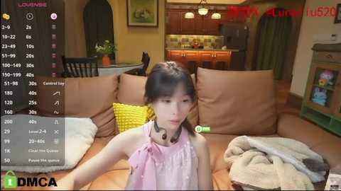 Media: A video of an East Asian woman with straight black hair, wearing a pink sleeveless dress, sitting on a brown couch in a cozy living room, with a kitchen visible in the background.