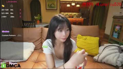 Media: A video captures an Asian woman with long black hair, wearing a white top, sitting on a brown leather couch in a cozy living room.