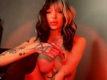 Media: Video of a topless woman with long, wavy hair, dark and light highlights, tattoos on her arms, wearing red makeup, with a red-lit background.