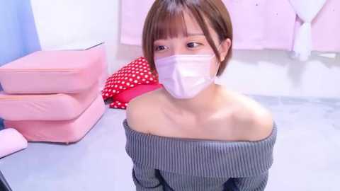 Media: Video of an Asian woman with short brown hair, wearing a gray ribbed sweater, off-shoulder, and a white face mask, kneeling in a brightly lit room with pink and blue furniture, polka-dot dress, and a white glove.