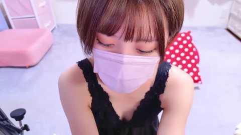 Media: A video of a young Asian woman with short brown hair, wearing a black lace top and a pink face mask, seated indoors on a light blue floor, with a red polka-dot cushion and pink storage boxes in the background.