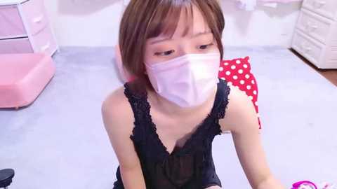 Media: Video of an Asian woman in a surgical mask, black lace lingerie, kneeling in a bright, clean room with pink furniture and white cabinets.