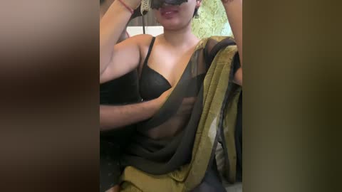 Media: Video of a woman wearing a black and green saree, with a black bra underneath, holding a green shawl, blindfolded, partially obscured by a blurred brown object.